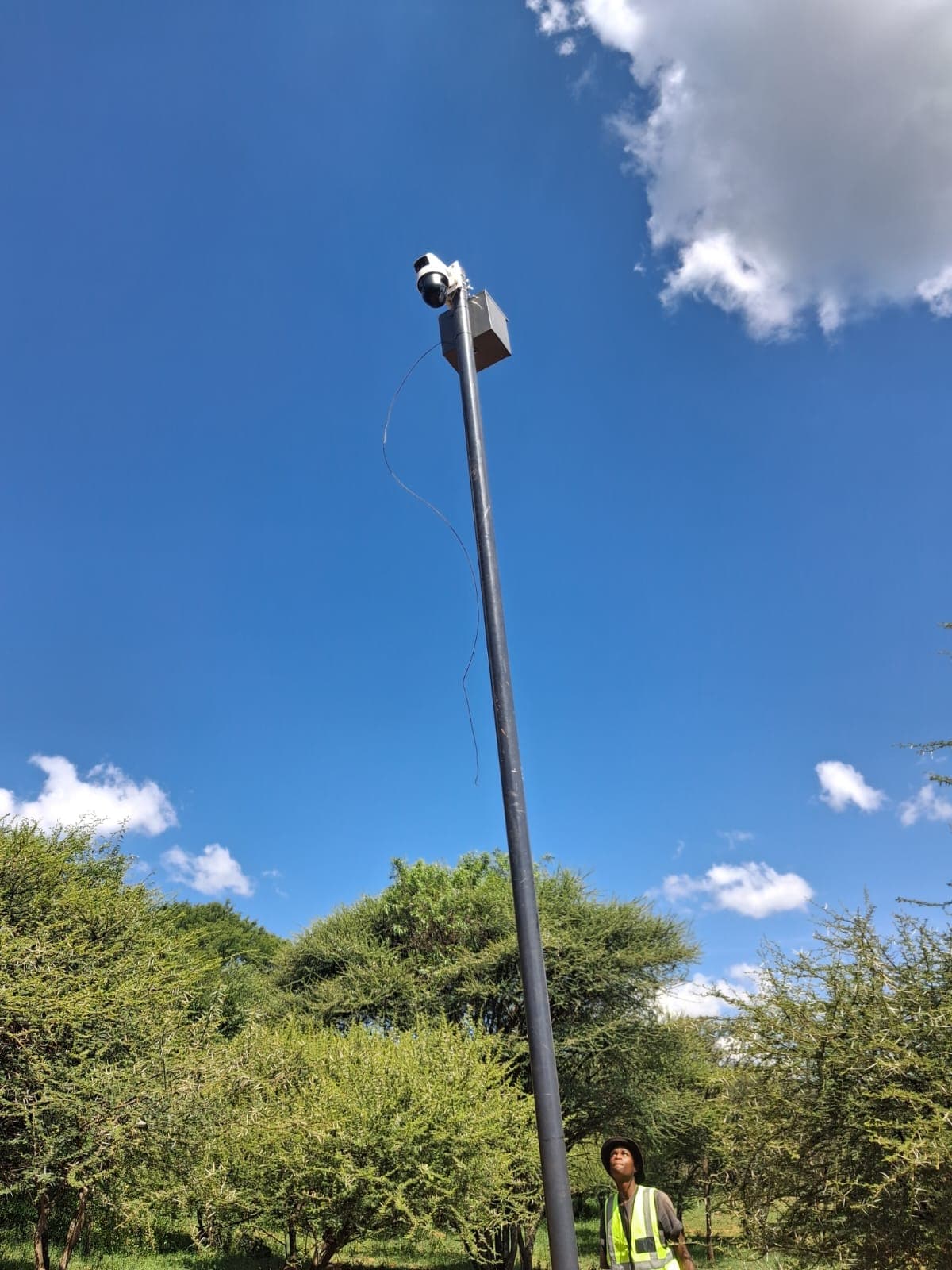 Security camera and control box atop a tall pole against a bright blue sky.