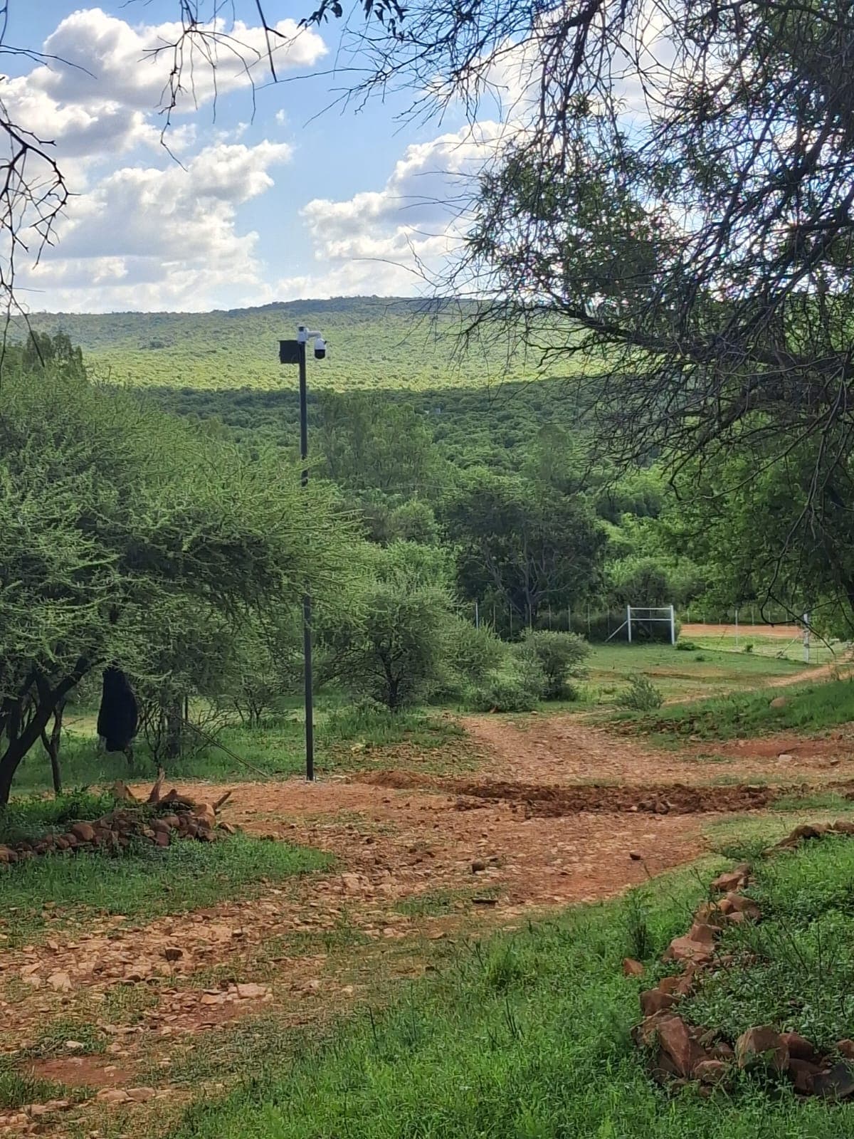 Security camera pole overlooks a dirt path winding through lush green hills under a bright sky.