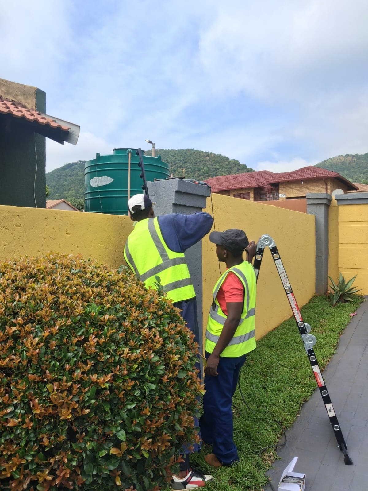 Two workers in high-visibility vests install equipment on a yellow wall near a water tank.