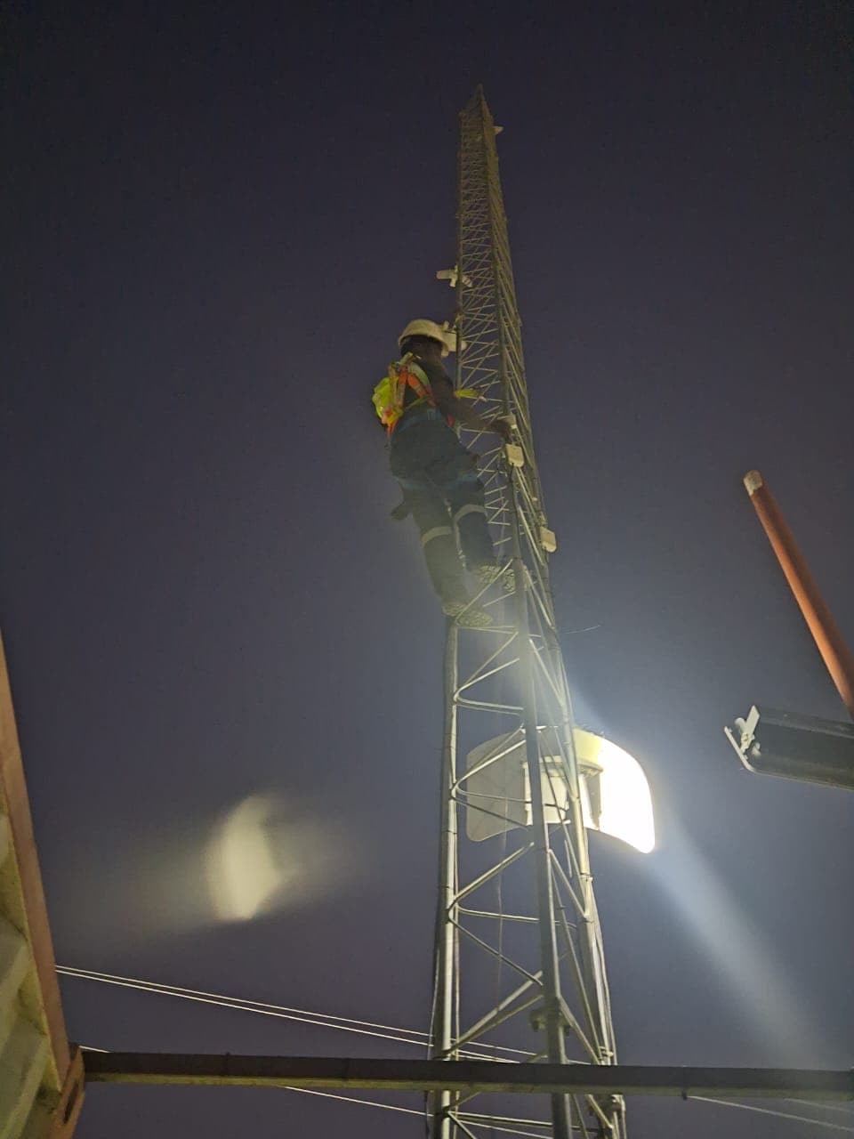 Worker in safety gear climbs a tall metal lattice tower at night under bright lights.
