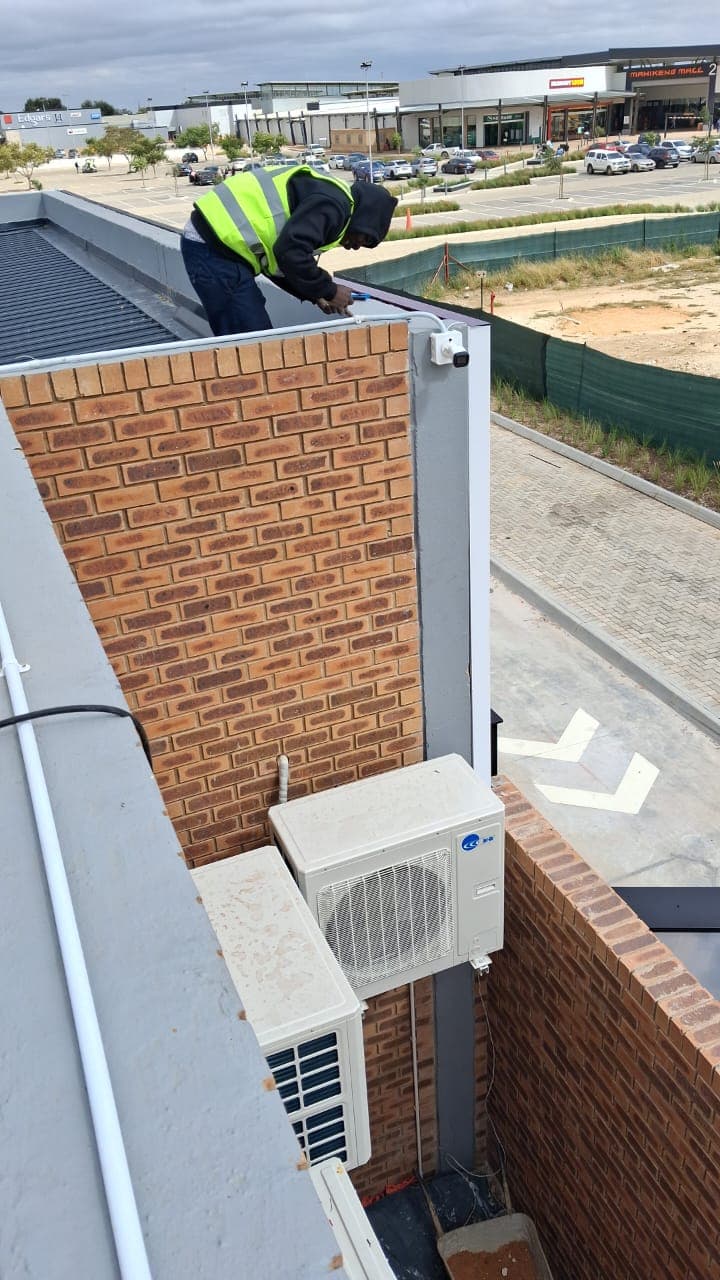 Technician in a high-visibility vest installs a security camera on a brick building's roof.