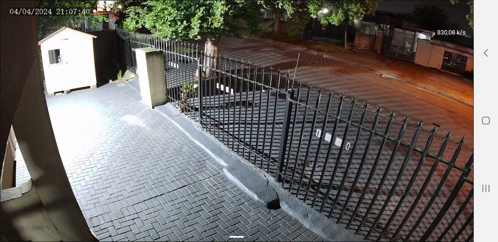 Nighttime security footage showing a grey brick driveway, black spiked fence, and small white shed.