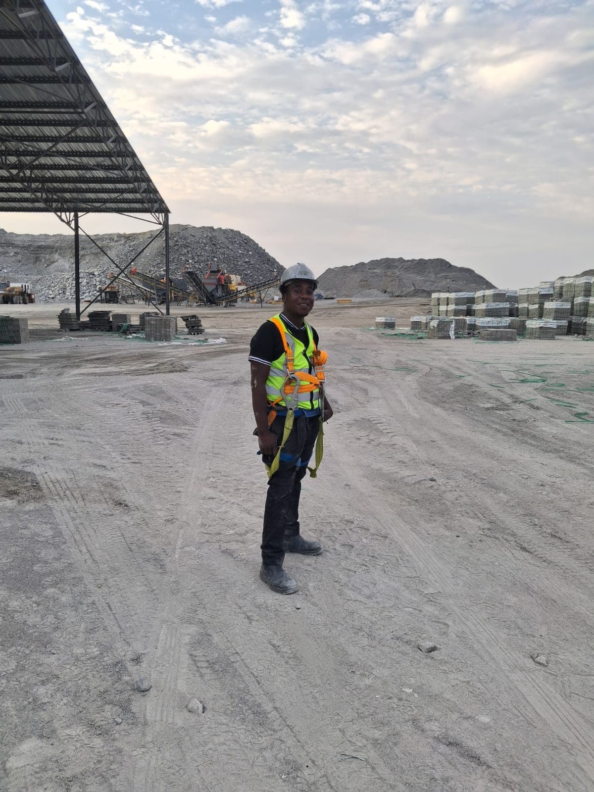 Construction worker in safety gear stands in a dusty quarry with gravel piles and equipment.