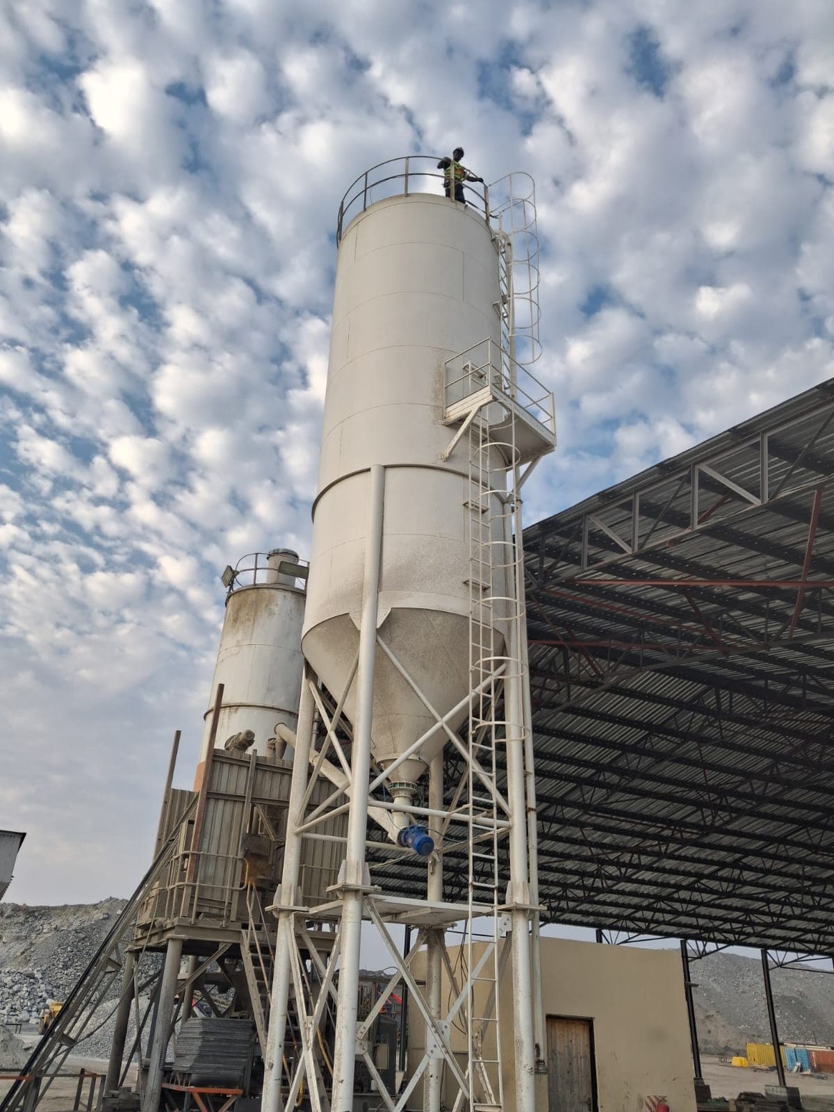 Tall white industrial silo with a worker on top, set against a blue, cloudy sky.