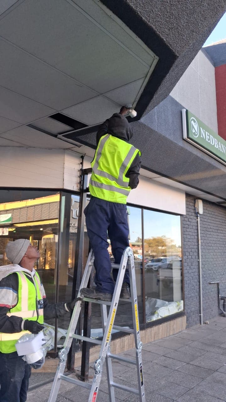 Two workers in safety vests install a security camera on a building's outdoor ceiling.