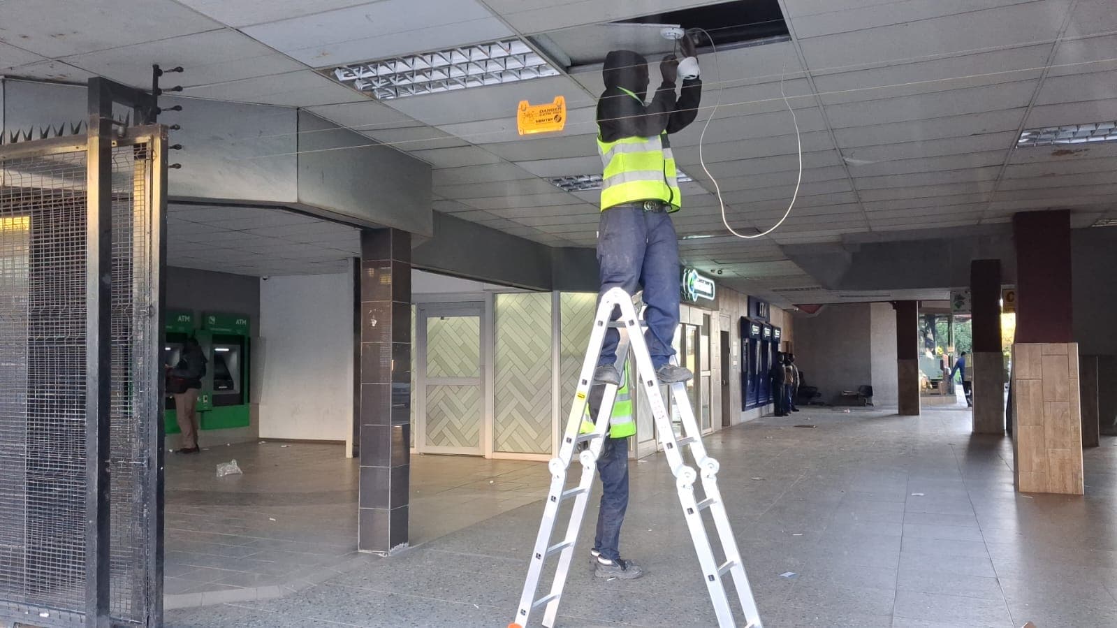 Worker in a safety vest on a ladder repairing a drop ceiling in a hallway.