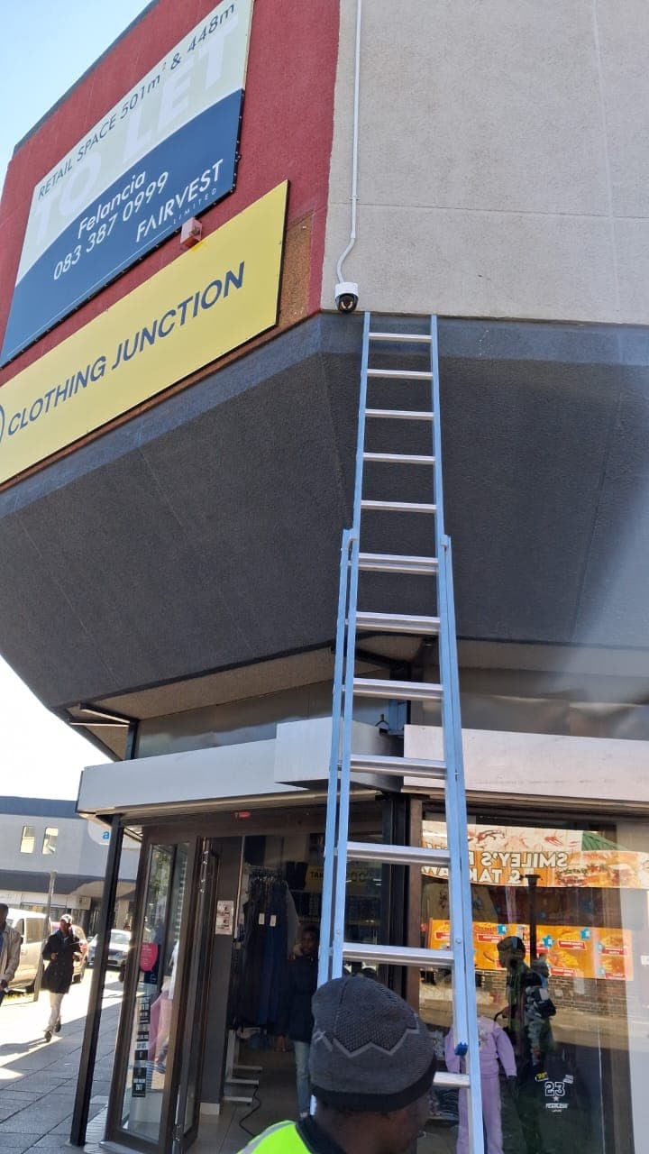 Silver ladder leaning against a building corner near a security camera and Clothing Junction sign.