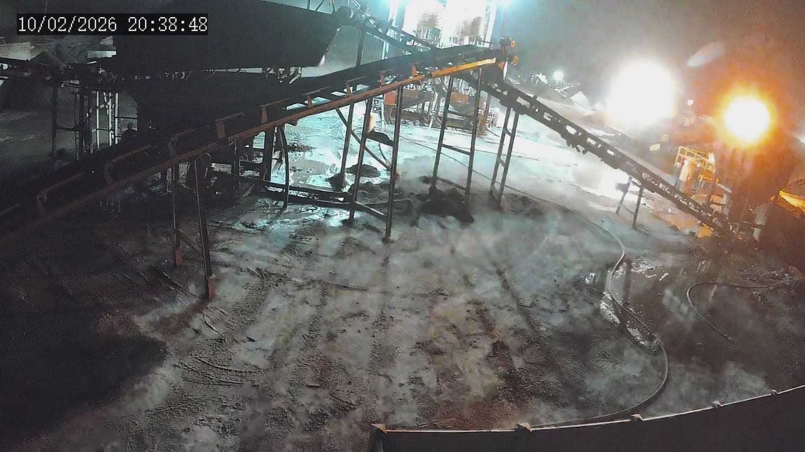 Industrial conveyor belt system at night with bright lights illuminating muddy ground and mist.