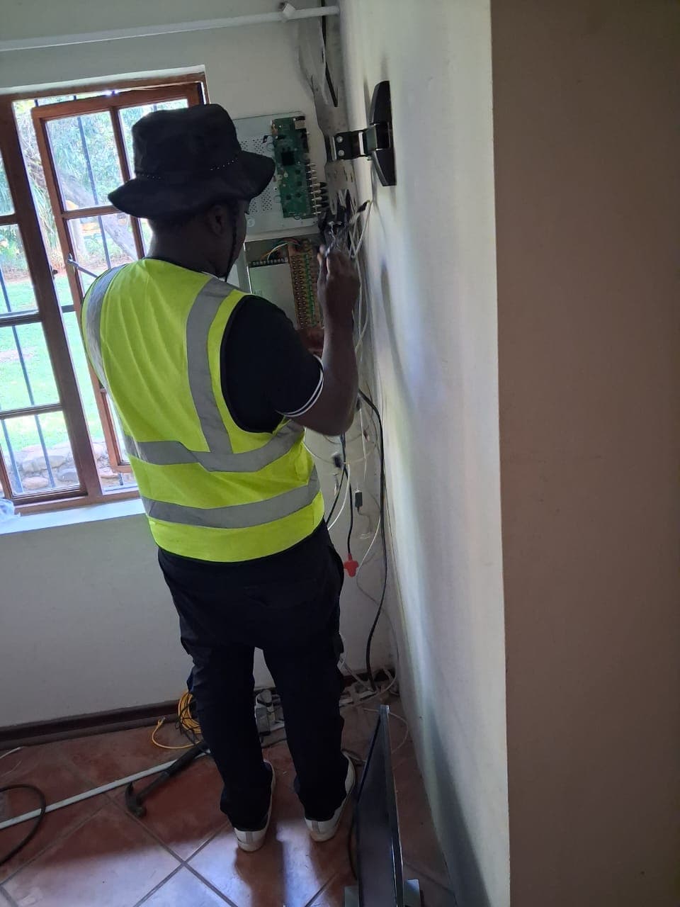 Technician in a yellow safety vest works on electrical wiring and circuit boards on a wall.